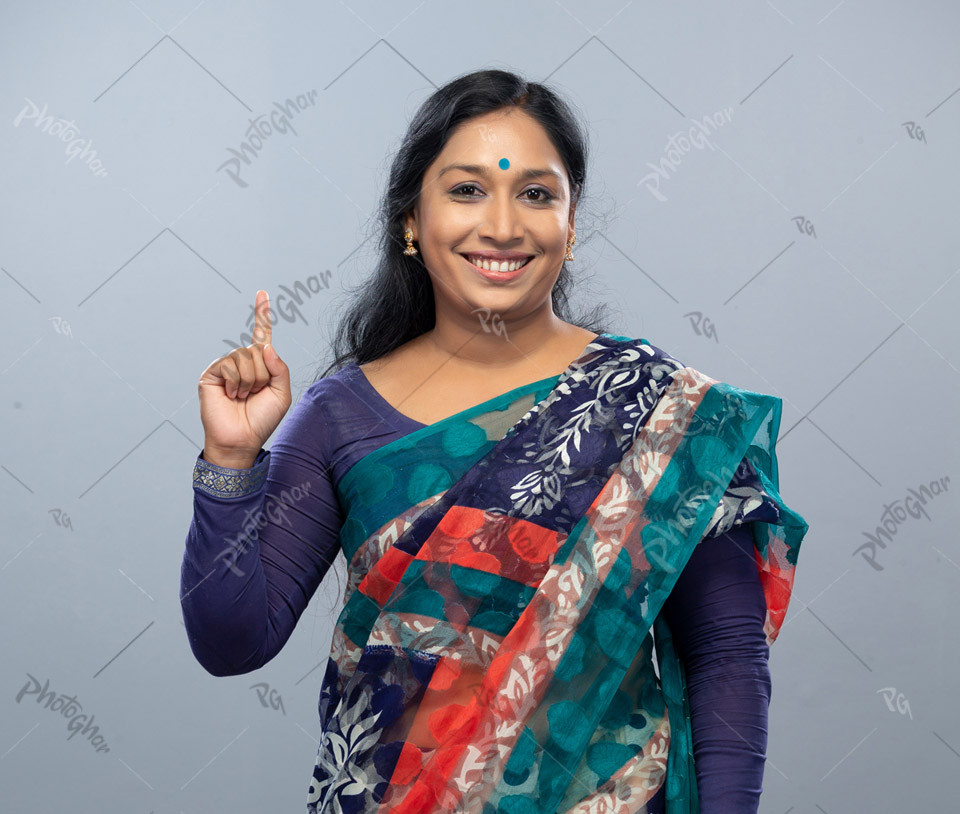 Close-up view of a beautiful housewife in traditional saree with straight black hair, pointing up to the empty copy space for advertisement, standing confidently, looking happily, against a gray studio backdrop.